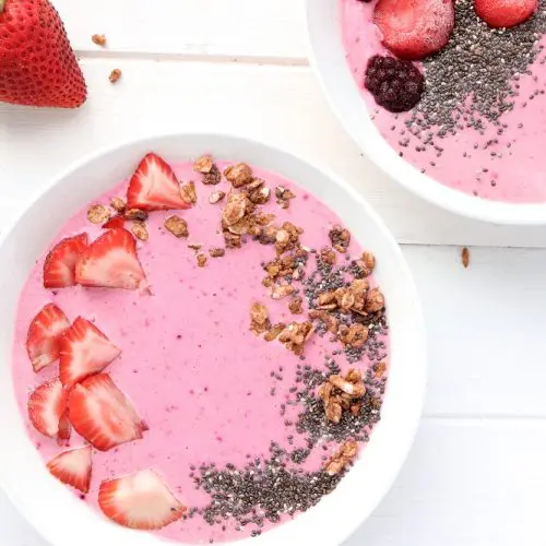 Two white bowls holding pink colored kefir garnished with strawberries, chia seeds and granola