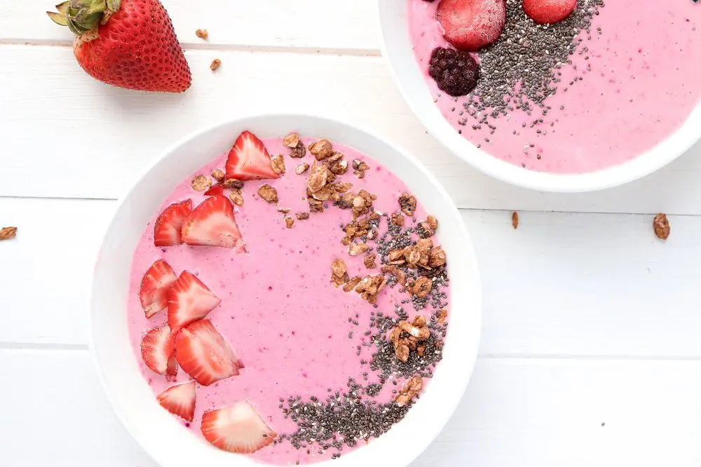 Two white bowls holding pink colored kefir garnished with strawberries, chia seeds and granola