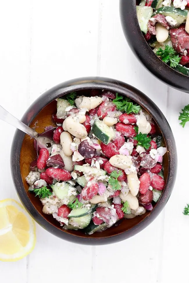 Two small wooden bowls of beans, cucumber and olives garnished with parsley and a lemon wedge