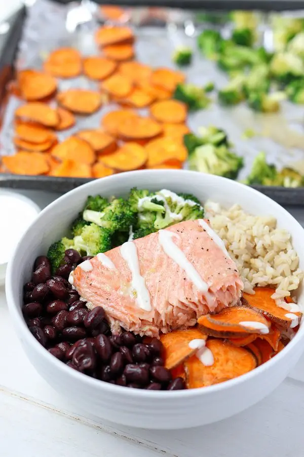 salmon buddha bowl drizzled in maple tahini dressing in front of sheet pan of extra veggies 