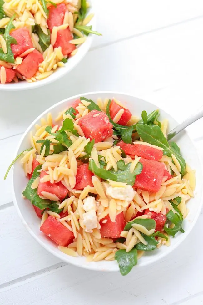 spoon in a bowl of orzo salad with chunks of watermelon